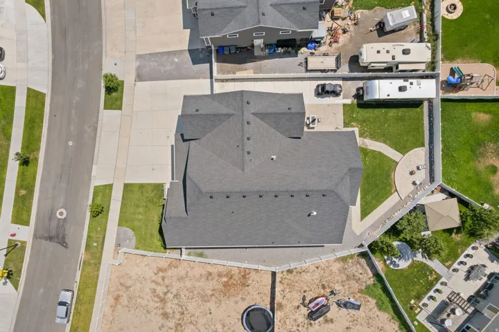 Aerial view showcasing a dark shingle roof, concrete driveway, and surrounding green lawn