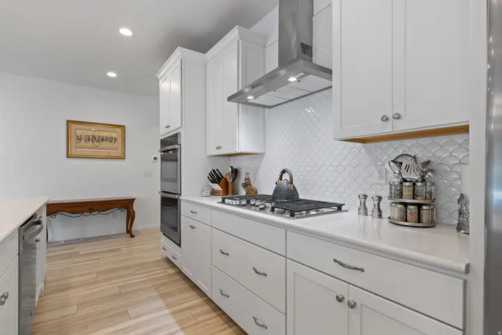 Kitchen featuring white cabinetry, light-toned wood-finish flooring, a stainless steel range hood, double wall ovens, and a scallop-patterned white tile backsplash