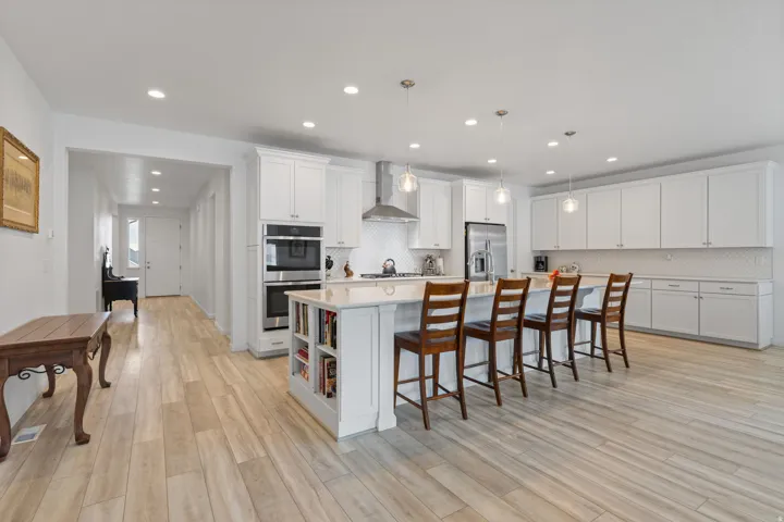 Bright kitchen featuring a substantial island with integrated shelving, pendant lighting, white cabinetry, a built-in double oven, and wood-finish flooring