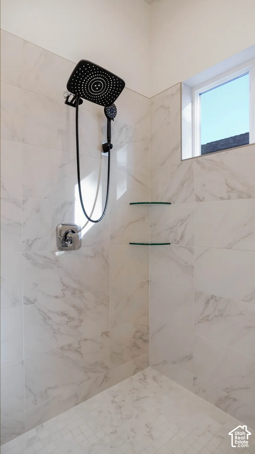 Walk-in shower featuring large format white tile walls with grey veining, a black multi-function showerhead with handheld sprayer, chrome fixtures, two clear glass corner shelves, and a square window