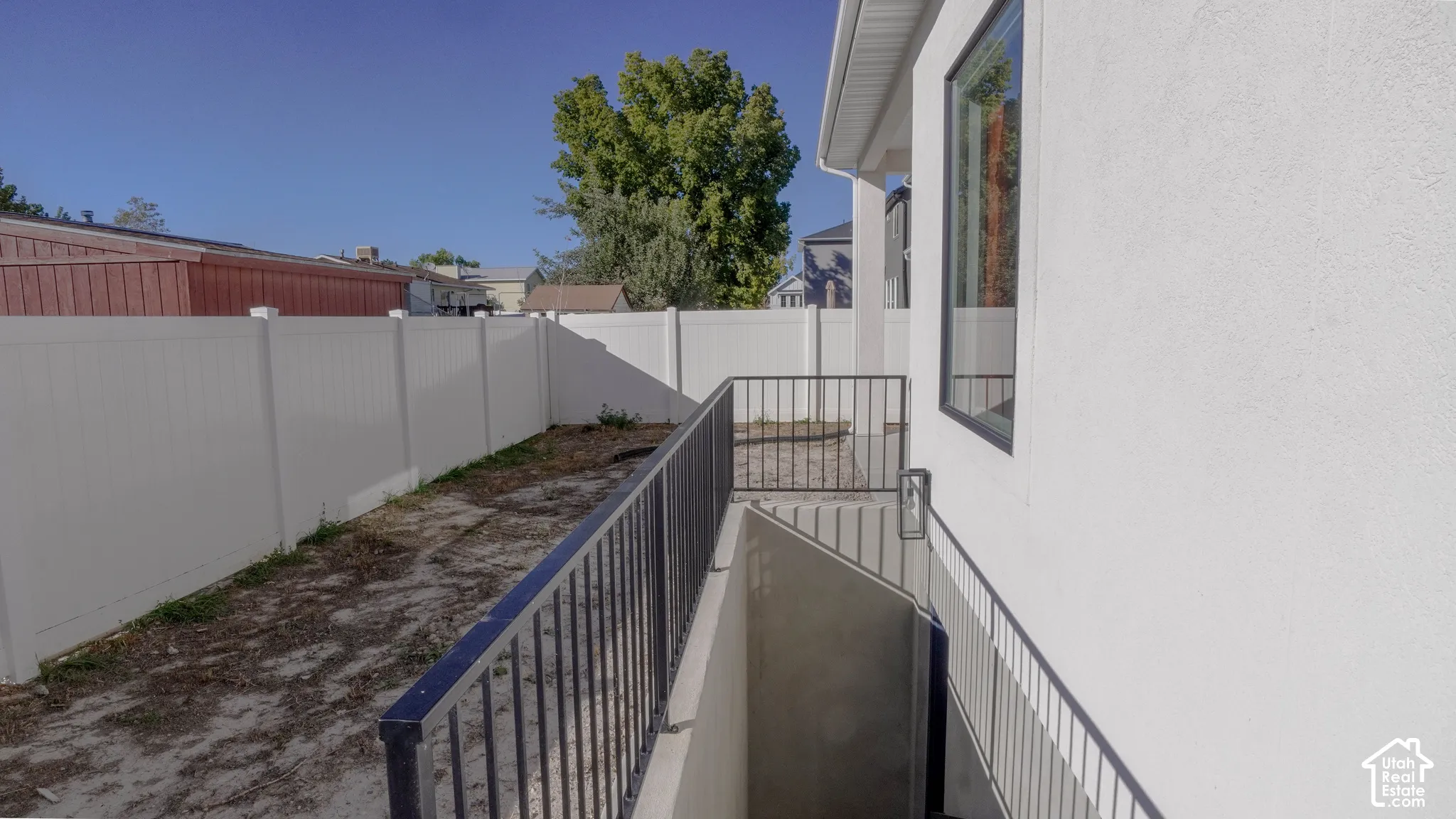 Exterior stucco siding with a black-framed window, complemented by a white privacy fence and a dark metal railing leading to a concrete basement entry