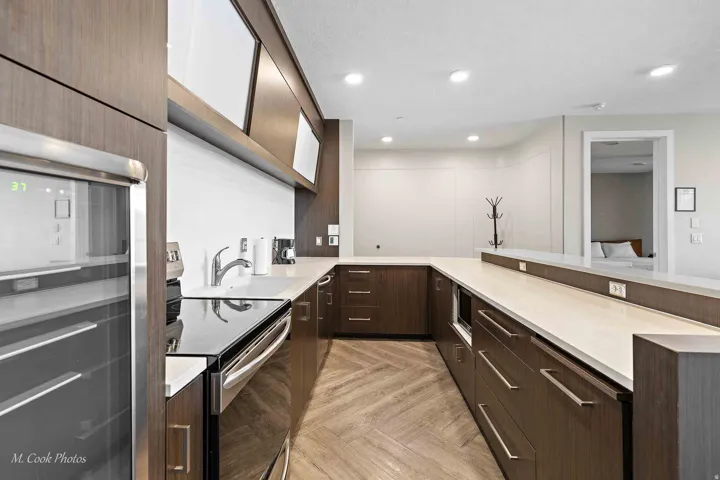 Modern kitchen featuring dark wood cabinetry with sleek metal hardware, dual undermount sinks with chrome fixtures, and quartz countertops spanning the galley layout