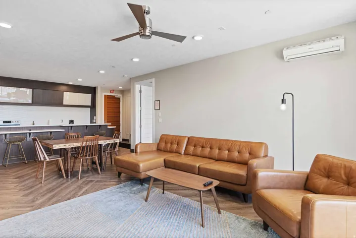 Open-concept living space featuring a leather sectional sofa and matching accent chair, hardwood-finish flooring, and abundant natural light from recessed ceiling fixtures and wall-mounted air conditioning unit
