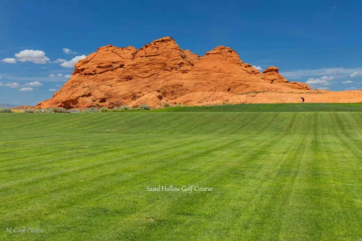 Expansive green golf course with striped fairways, framed by striking red rock formations under a clear blue sky