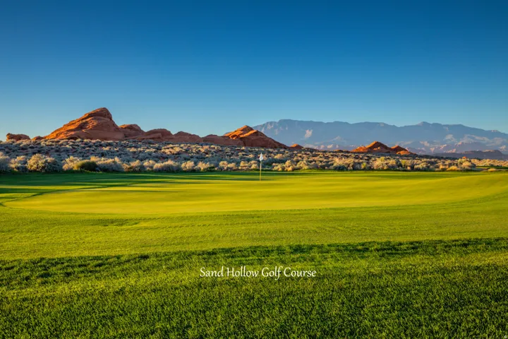 Expansive golf course green with a flagstick, bordered by arid shrubbery and striking red rock formations