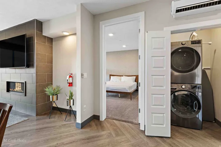Open-concept living space featuring herringbone wood-finish flooring, a linear gas fireplace with tile surround and built-in shelving, wall-mounted flat-screen television, and direct access to a laundry room equipped with stacked front-load washers and dr