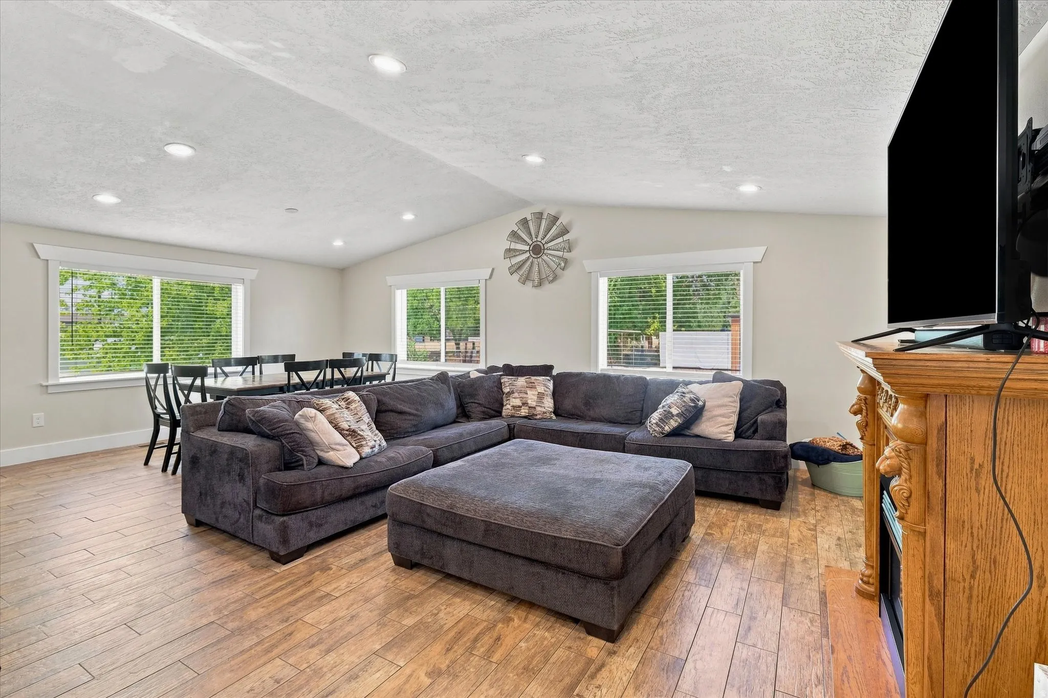 Spacious open-concept living area featuring wood-finish flooring, a vaulted ceiling with recessed lighting, and multiple windows with white trim