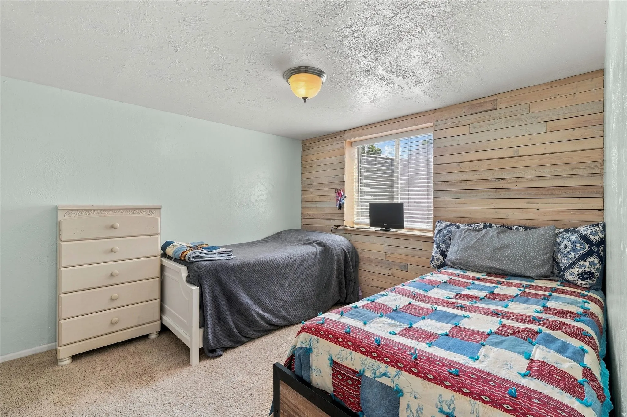 Comfortable bedroom featuring dual beds with coordinating bedding, wood-finish accent wall, carpeted flooring, four-drawer dresser with ample storage, and recessed ceiling lighting