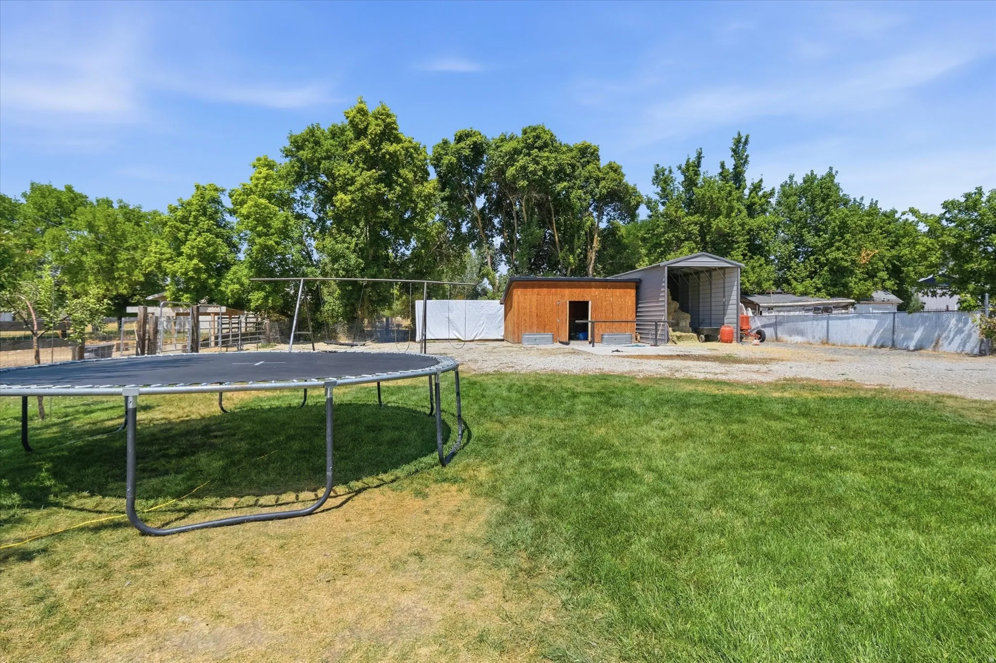 Spacious recreational property featuring a large manicured lawn with mature shade trees, a trampoline, and multiple outbuildings including an orange storage shed and metal structures