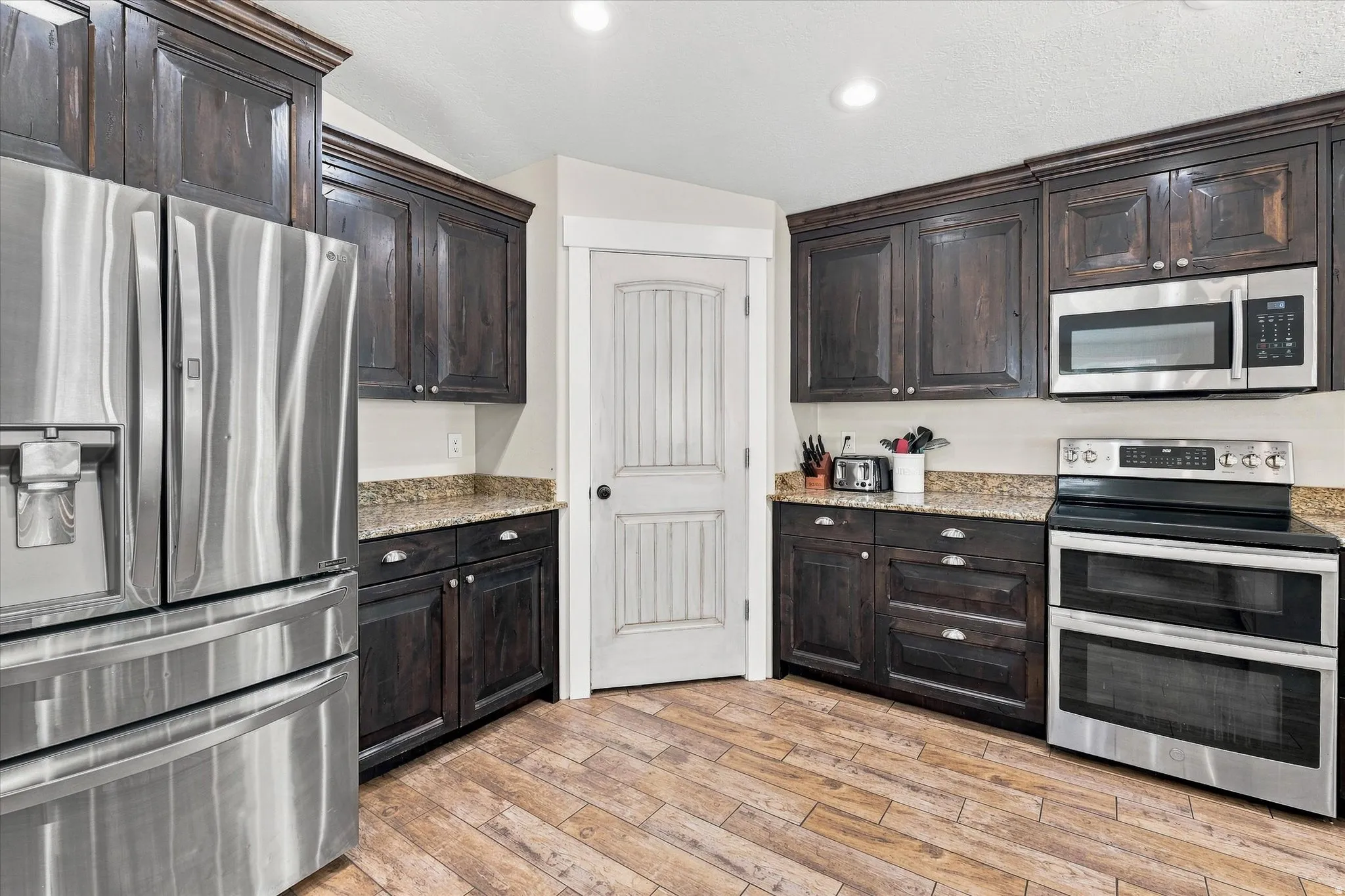 Elegant kitchen featuring dark cabinetry with glass-front uppers, stainless steel appliances including a French-door refrigerator, double wall ovens, and microwave, wood-finish flooring, granite countertops, and recessed lighting throughout