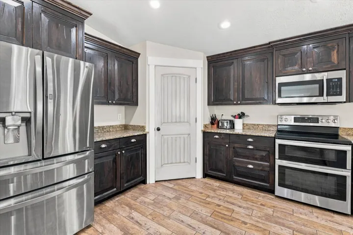 Elegant kitchen featuring dark cabinetry with glass-front uppers, stainless steel appliances including a French-door refrigerator, double wall ovens, and microwave, wood-finish flooring, granite countertops, and recessed lighting throughout