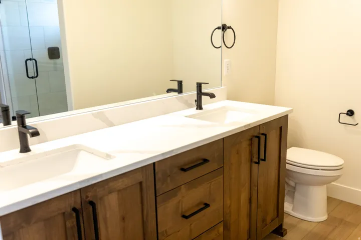 Dual vanity with integrated sinks and black fixtures, a spacious mirror, warm wood-finish cabinetry, and a walk-in shower with clear glass enclosure