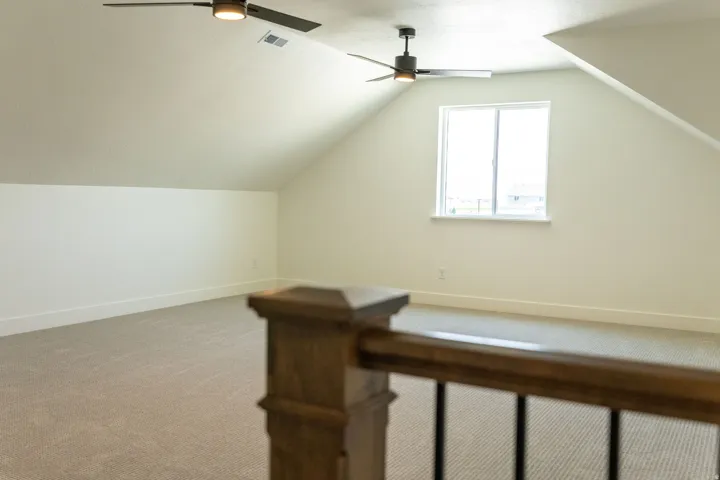 Carpeted bonus room with sloped ceilings, two ceiling fans with integrated lighting, and a window