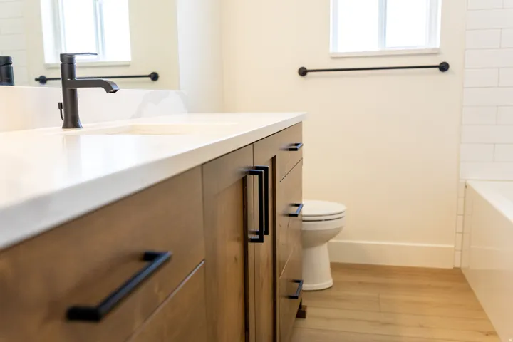 Wood-finish flooring, wood cabinetry with matte black hardware, white countertop with integrated sink, black single-handle faucet, and white subway tile surround