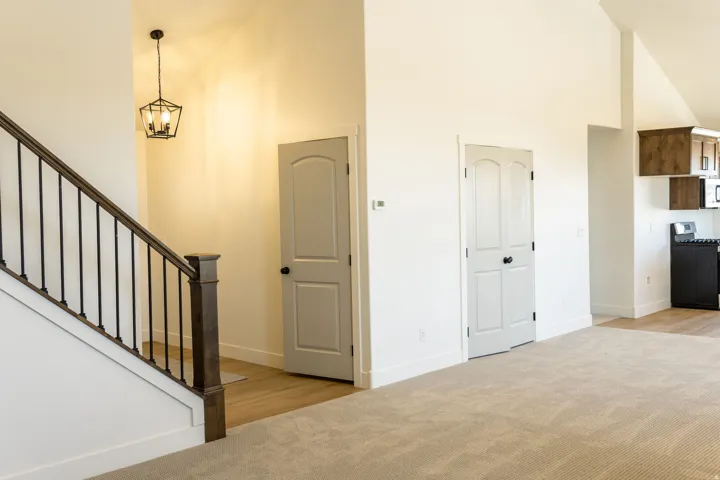 Open-concept living space with vaulted ceilings, a staircase featuring a dark wood handrail and black iron balusters, and light-colored carpeting