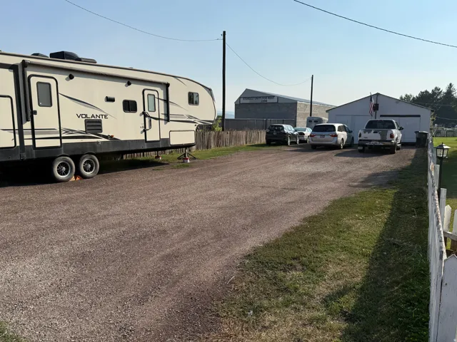 Spacious gravel lot with ample parking and vehicle storage, featuring a single-story recreational vehicle with slide-out sections and multiple outbuildings