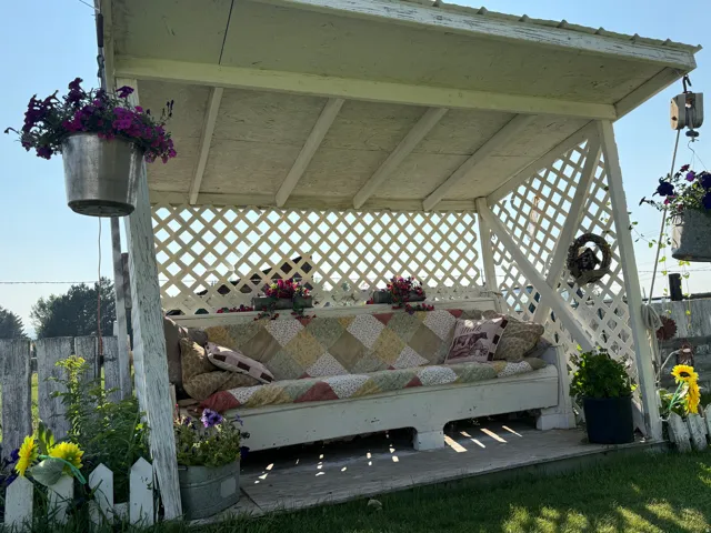 White lattice-sided gazebo with a built-in swing bench, featuring a white picket fence, and a green lawn