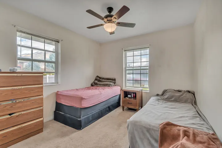 Carpeted bedroom featuring two windows with horizontal blinds, a ceiling fan with light fixture, and neutral-toned walls