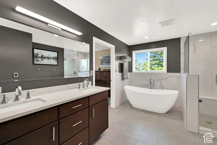Spa-inspired primary bathroom featuring dual vanities with wood-finish cabinetry and white quartz countertops, a freestanding soaking tub, walk-in shower with frameless glass enclosure and tile surround, linear ceiling lighting, and large windows