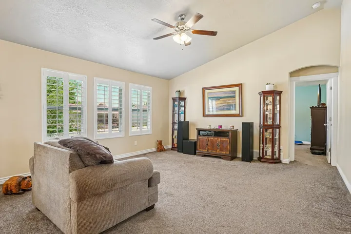 Vaulted ceiling living area featuring three windows with plantation shutters, ceiling fan, carpet flooring, and an arched doorway