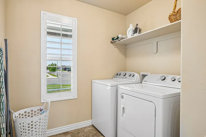 Dedicated laundry room featuring stacked front-load washer and electric dryer with overhead shelving for storage, tile flooring, and natural light from a large window with plantation shutters