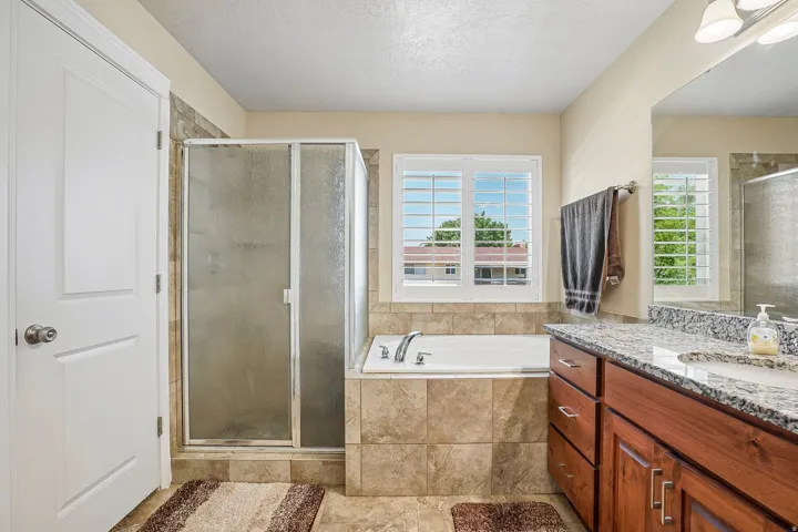 Bathroom featuring a walk-in shower with glass enclosure, a built-in tub, dual-pane window with white plantation shutters, a wood-finish vanity with an integrated sink, and stone-finish countertops