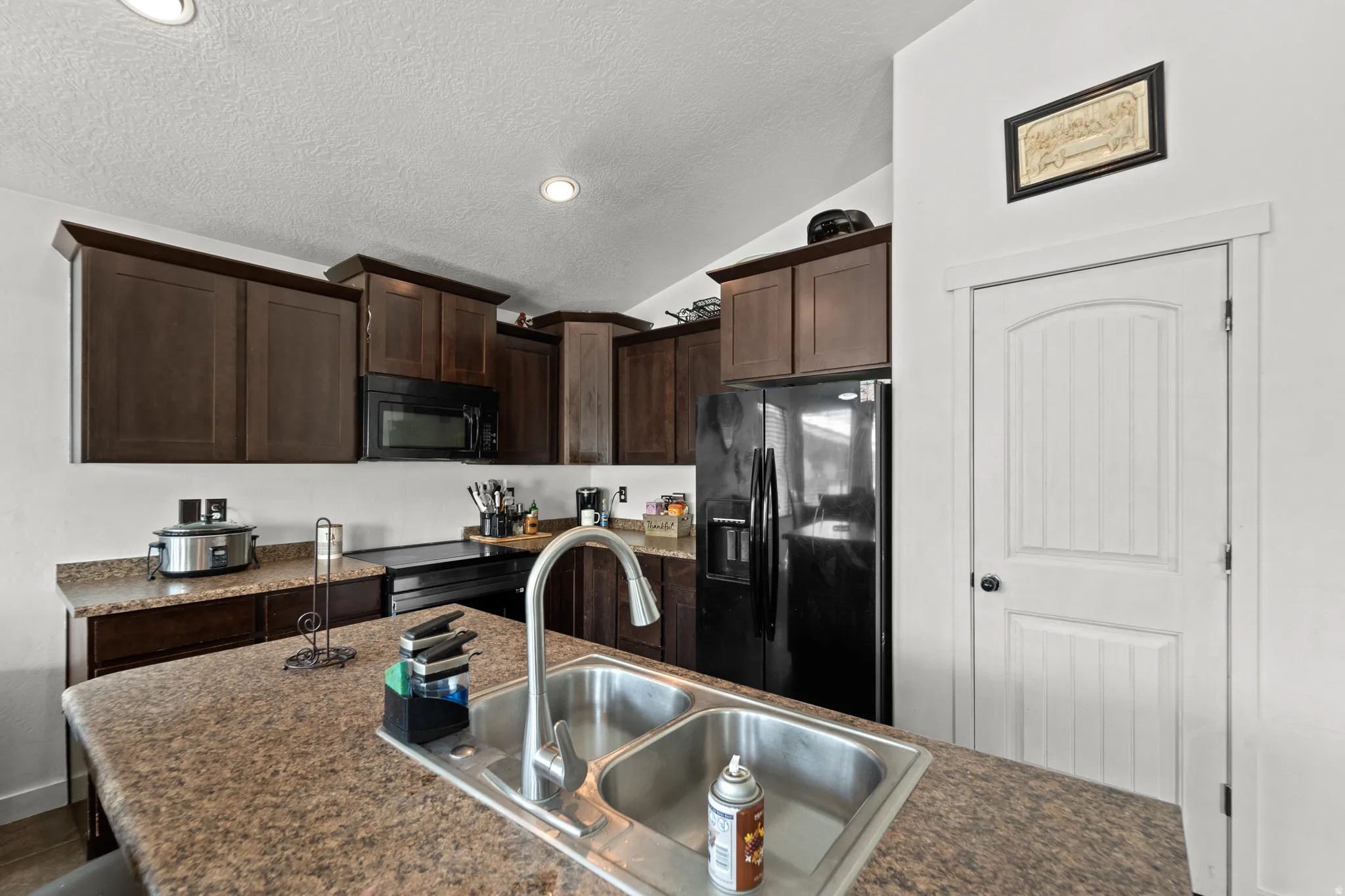 Kitchen featuring a dual-basin stainless steel sink, dark wood-finish cabinetry, black appliances, recessed lighting, and a textured countertop