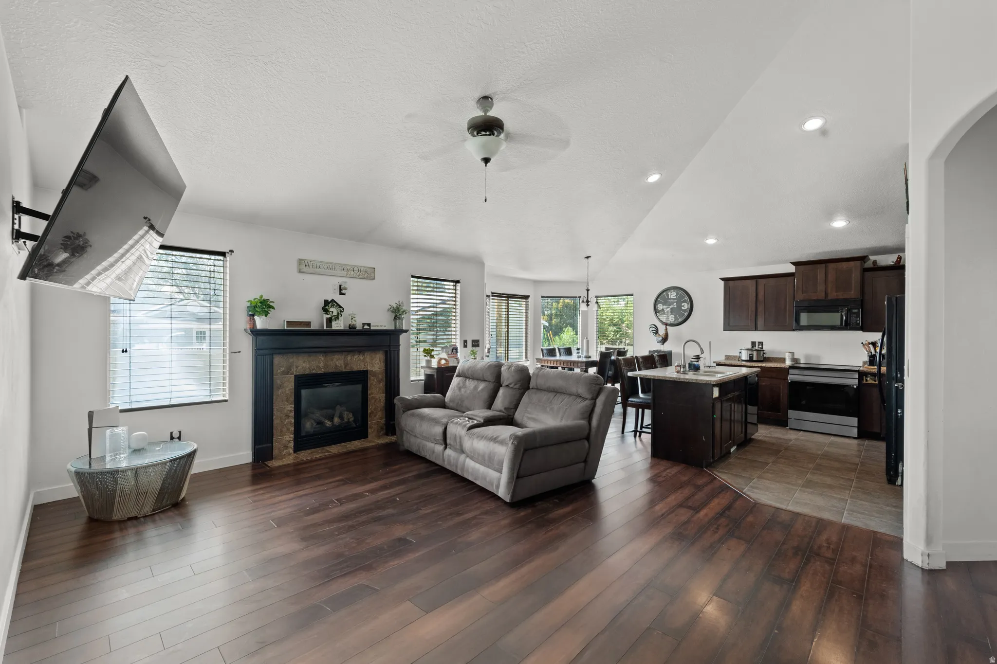 Spacious living area featuring rich wood-finish flooring, a built-in fireplace with a dark mantel and stone surround, and an open layout to the kitchen