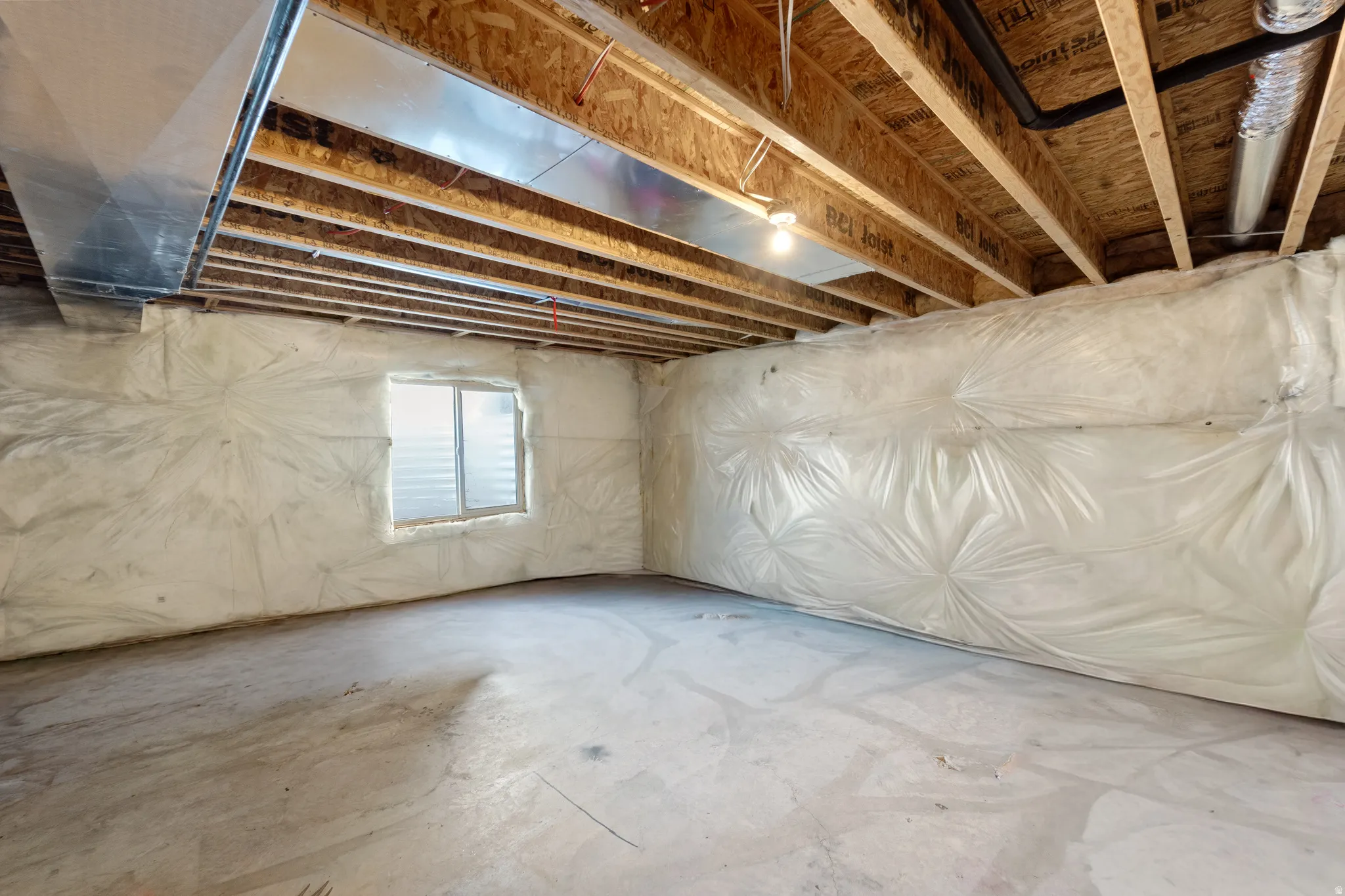 Unfinished basement space featuring exposed wood joists, concrete flooring, foundational insulation, and a single window with horizontal blinds