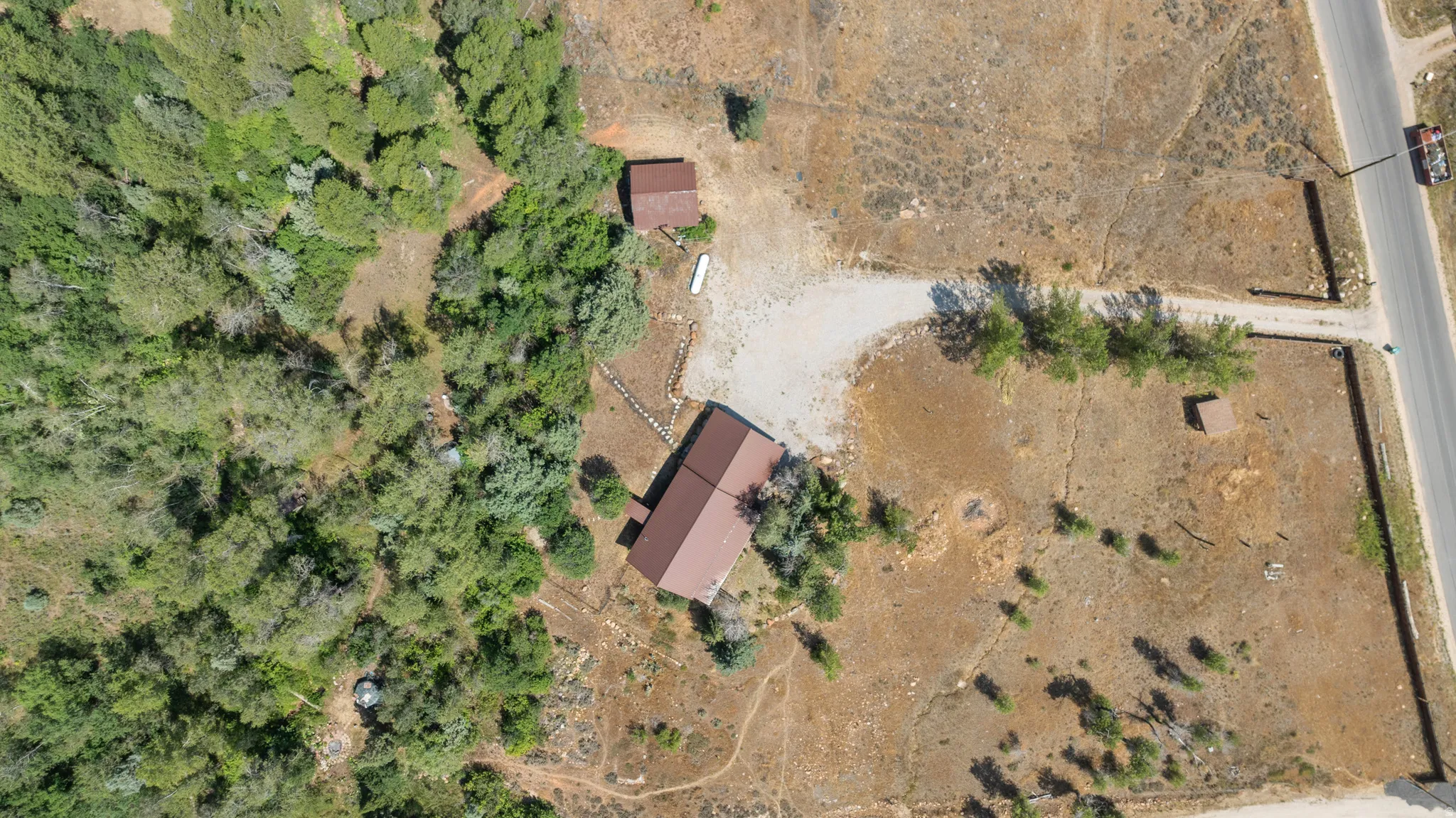 Extensive property featuring a primary structure with a brown roof, a smaller accessory building, a gravel driveway, and a partially wooded landscape