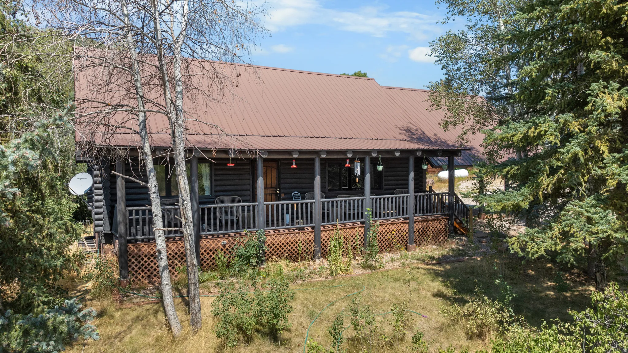 Log home exterior featuring a brown metal roof, full-length covered porch with dark railing, and natural wood lattice skirting