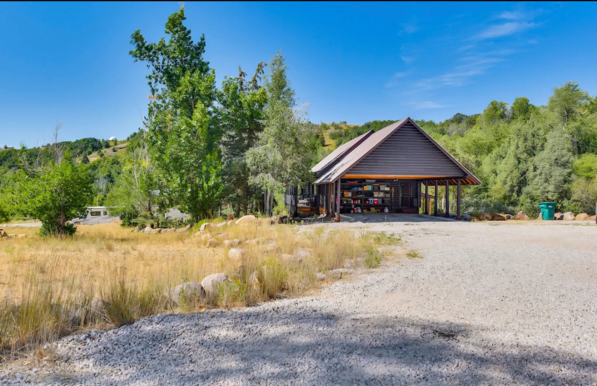Wood-sided structure featuring an open carport, gabled roof, and gravel driveway