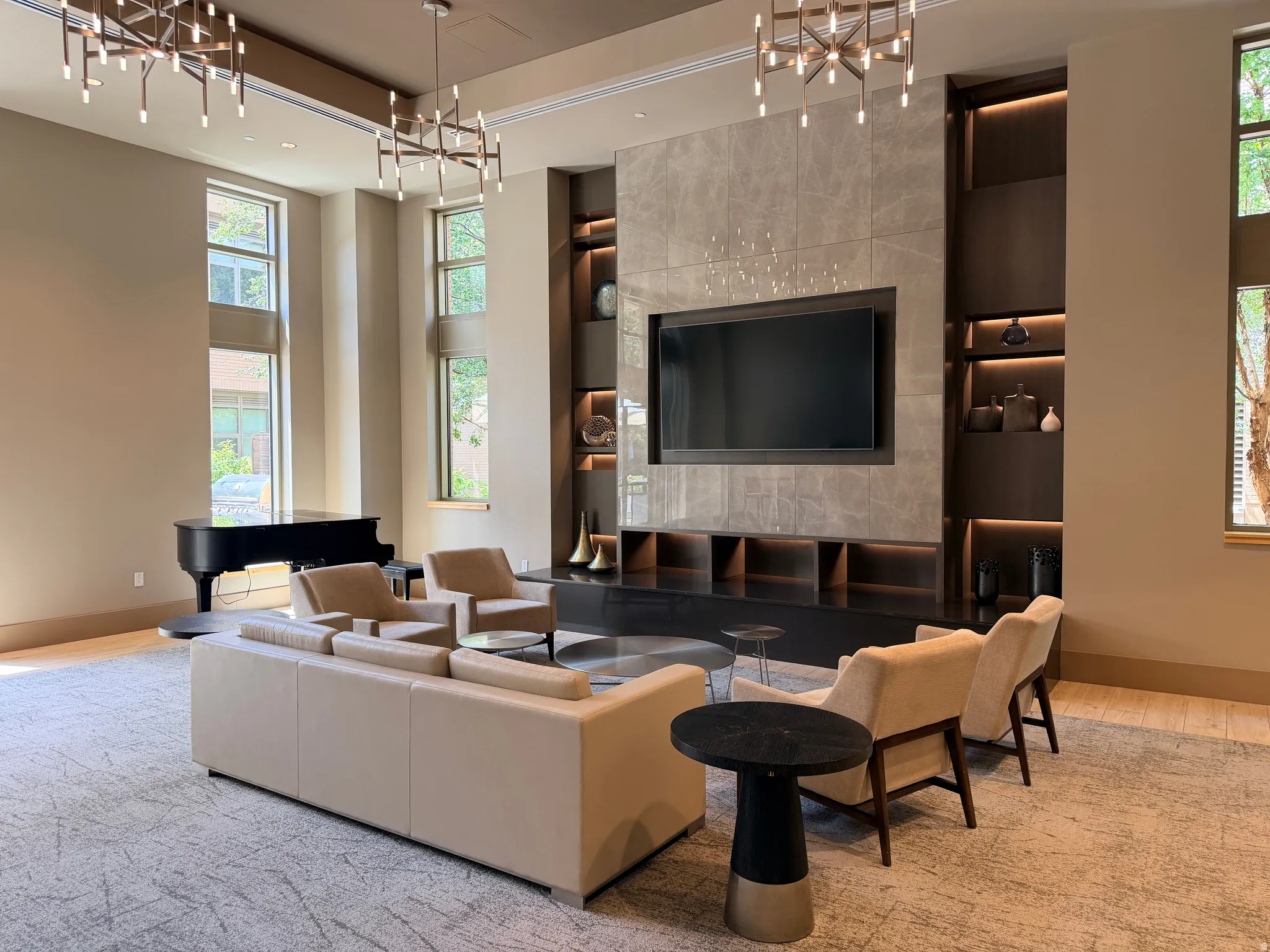 Spacious living area featuring floor-to-ceiling windows, two contemporary chandeliers, a built-in media wall with integrated shelving, and patterned area carpeting