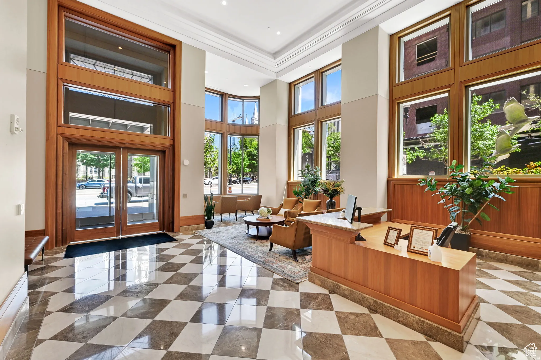 Grand entryway featuring a checkerboard-patterned floor, expansive wood-framed windows, a double-door glass entrance, a built-in wood reception desk, and high ceilings with recessed lighting