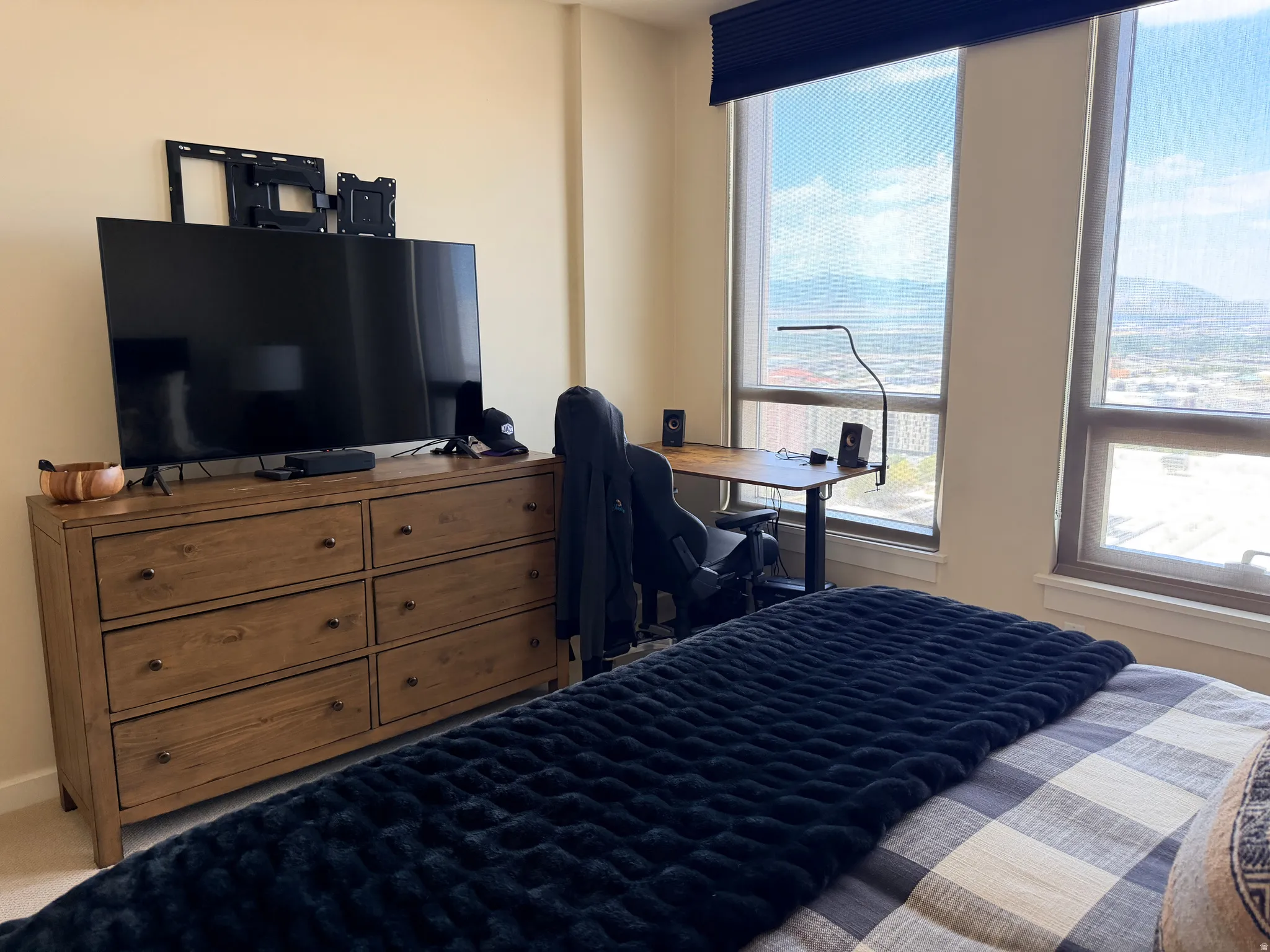 Bedroom featuring expansive windows with panoramic vistas, wood-finish dresser with multiple drawers and integrated storage shelving, flat-screen television mounted above, dedicated desk workspace with ergonomic seating, and plush bedding with coordinatin