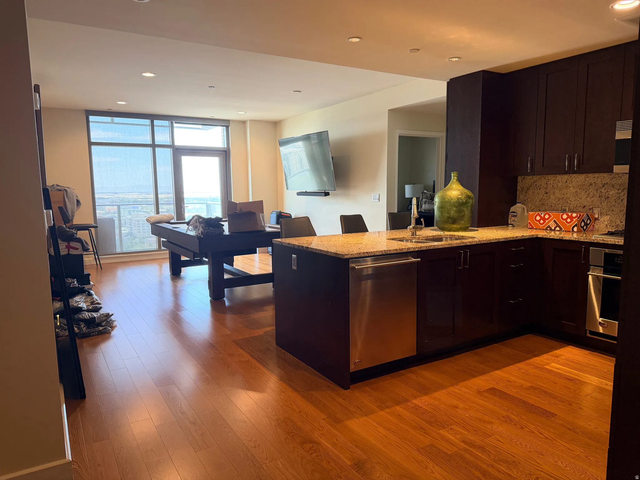 Open-concept kitchen and living area featuring custom dark cabinetry with stainless steel appliances, wood-finish flooring throughout, a large center island with breakfast bar seating, recessed ceiling lighting, and expansive windows with natural light an