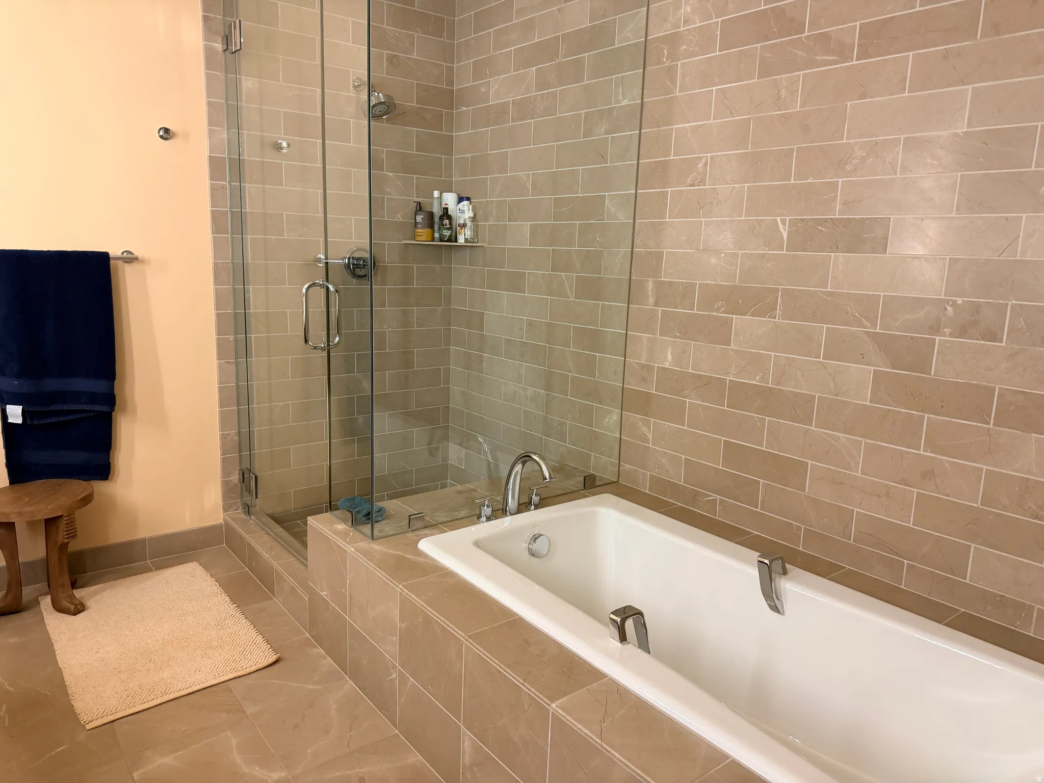 Spa-inspired primary bathroom featuring a frameless glass walk-in shower enclosure with subway tile surround, a soaking tub with deck-mounted faucet, heated towel rack, recessed shelving, and neutral stone-finish flooring throughout