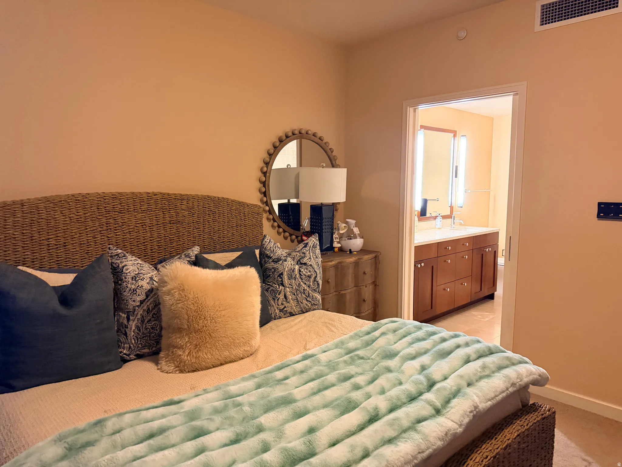 Bedroom featuring neutral wall color, carpet flooring, a built-in air vent, and an open doorway revealing a bathroom with a dual vanity