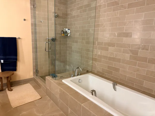 Spa-inspired primary bathroom featuring a frameless glass walk-in shower enclosure with subway tile surround, a soaking tub with deck-mounted faucet, heated towel rack, recessed shelving, and neutral stone-finish flooring throughout