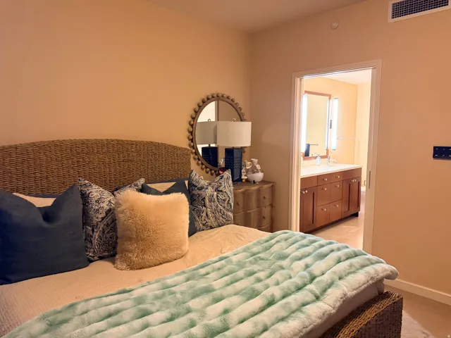 Bedroom featuring neutral wall color, carpet flooring, a built-in air vent, and an open doorway revealing a bathroom with a dual vanity