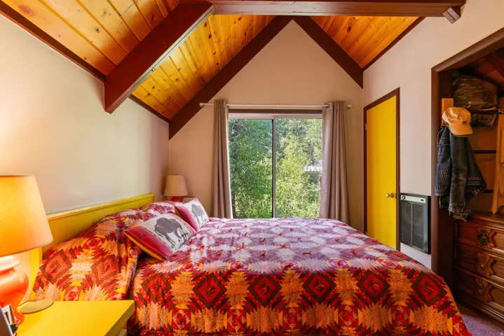 Bright primary bedroom featuring exposed wood beam ceiling, built-in yellow headboard, dual glass doors opening to natural landscape, wood-finish flooring, and recessed lighting throughout