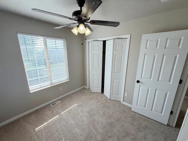 Neutral wall paint, carpet flooring, a window with horizontal blinds, a ceiling fan with light fixture, and white paneled doors