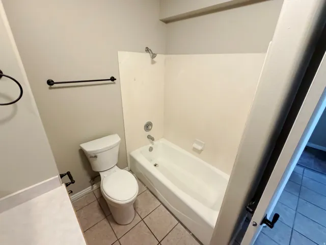 Bathroom featuring a white bathtub with shower, white toilet, and beige tile flooring