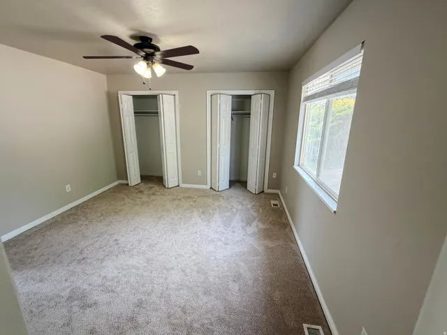 Carpeted room featuring a ceiling fan with integrated lighting, two bi-fold door closets, and a large window