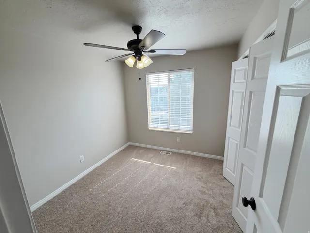 Neutral-toned wall paint, textured carpeting, window with blinds, ceiling fan with integrated lighting, and white paneled doors