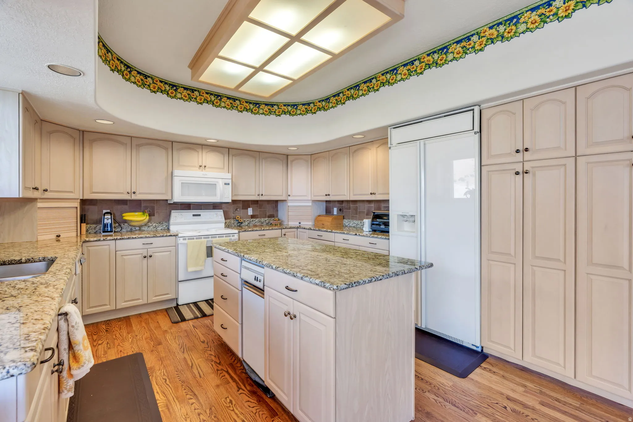 Spacious kitchen featuring light-toned cabinetry, granite-finish countertops, wood-finish flooring, a built-in kitchen island with integrated dishwasher, and a large recessed ceiling light fixture
