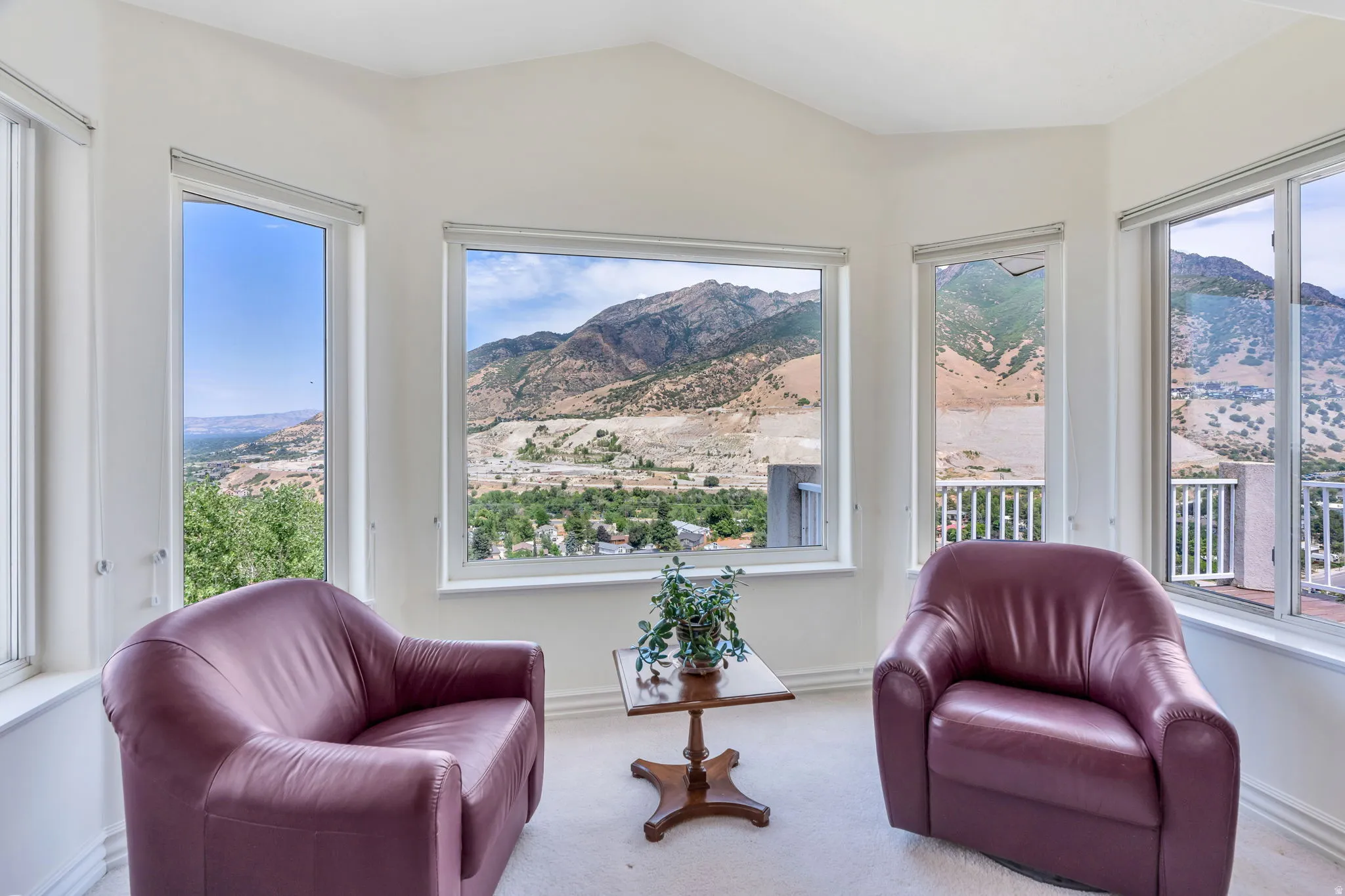 Sunlit master room showcasing expansive multi-panel windows framing mountain and valley vistas, burgundy leather armchairs, neutral carpet flooring, and abundant natural light throughout the space
