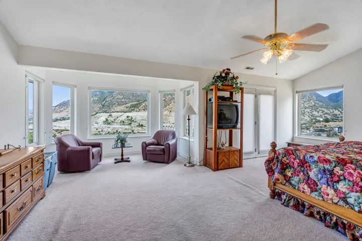 Spacious primary suite with expansive windows framing mountain vistas, ceiling fan with integrated lighting, wood-finish dresser, and carpet flooring throughout