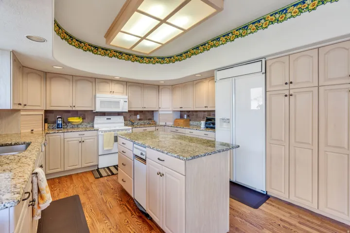 Spacious kitchen featuring light-toned cabinetry, granite-finish countertops, wood-finish flooring, a built-in kitchen island with integrated dishwasher, and a large recessed ceiling light fixture