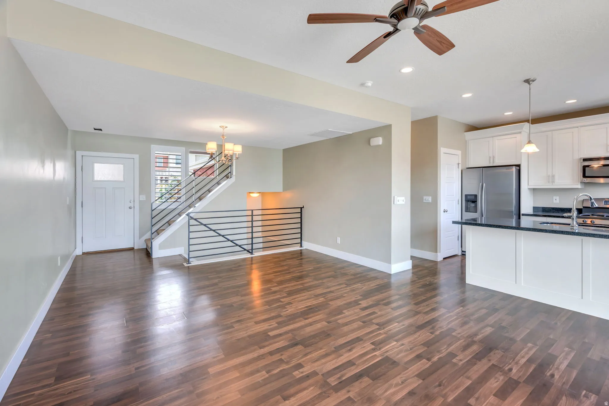 Open-concept living space featuring wood-finish flooring, a modern kitchen with white cabinetry and dark countertops, and a contemporary staircase with horizontal metal railings
